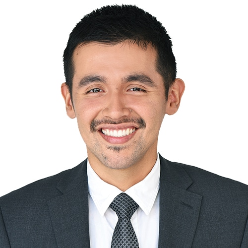 A professional portrait headshot of Jacob Hernandez, an associate attorney at Briones PC. He is smiling warmly and looking directly at the camera. He is wearing a dark charcoal grey suit jacket with a textured pattern, a crisp white dress shirt, and a dark, geometric-patterned tie. He has short, neat dark hair and a mustache. The background is a clean, neutral white studio backdrop.