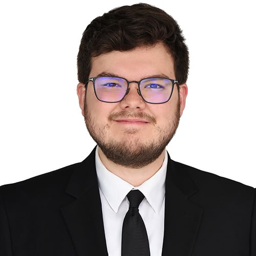 Professional headshot of Caleb Mead, Legal Analyst — a man with short brown hair, a beard and mustache, and glasses, wearing a black suit jacket, white dress shirt, and dark tie, against a plain white background.