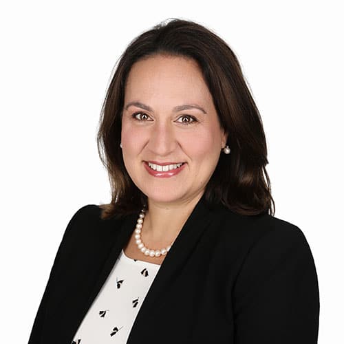 Studio portrait of Selen Perker smiling warmly at the camera. She has shoulder-length brown hair, brown eyes, and is wearing a black suit jacket over a white top with a subtle black print, paired with a pearl necklace and drop earrings.
